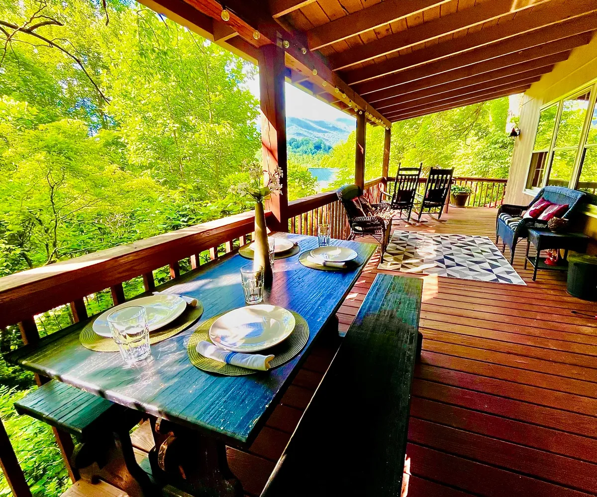 A vibrant exterior dining setup features a table set for a meal, cozy rocking chairs, and an expansive view of greenery and hills. The decorative rug and cheerful mood evoke the spirit of outdoor socializing and celebration.