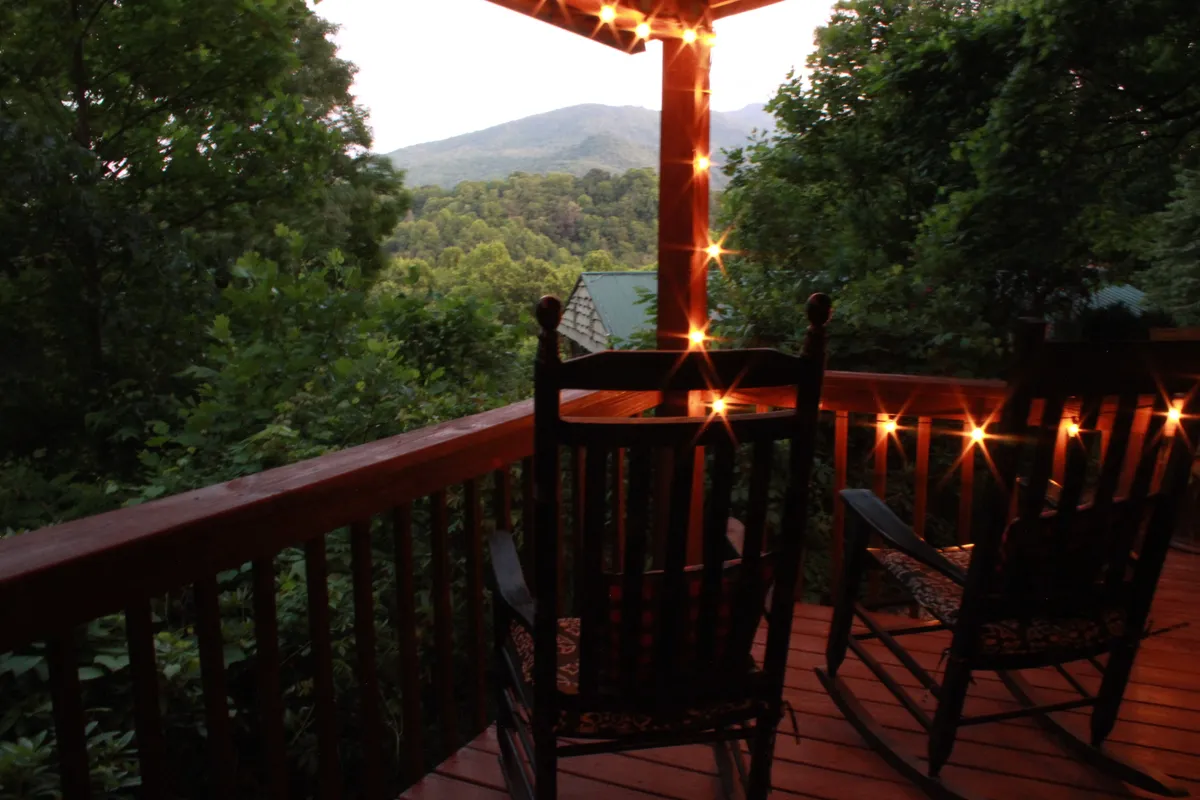 Two classic rocking chairs sit on a porch strung with twinkling lights, overlooking a breathtaking mountain vista. The scene evokes cozy evenings spent stargazing or enjoying a hot beverage, with the mountains providing a stunning, romantic backdrop.