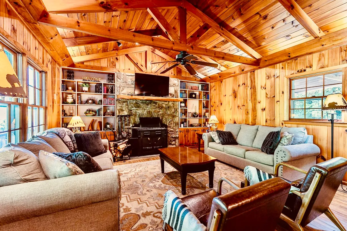 This image presents a central living room space featuring a dramatic stone fireplace flanked by bookshelves, comfortable seating, and classic wooden beams. Sunlight from large windows illuminates the heart of the gathering area, inviting relaxation and conversation.