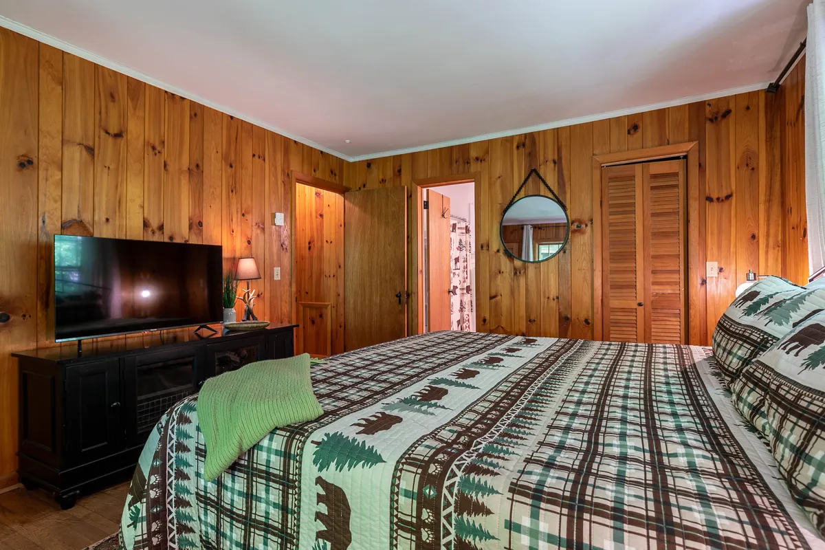 A comfortable bedroom with a patterned bedspread, a television, warm wooden walls, and a wall-mounted mirror. The setup invites relaxation, reading, or unwinding with a favorite show.