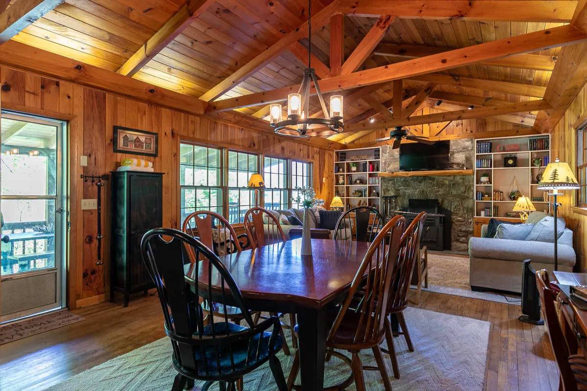 A wide, inviting shot of the open living and dining area, showing a spacious dining table, plush sofas, a fireplace, bookshelves, and expansive windows framing remarkable mountain views. The space glows with natural light and offers a seamless connection between the living, dining, and kitchen zones.