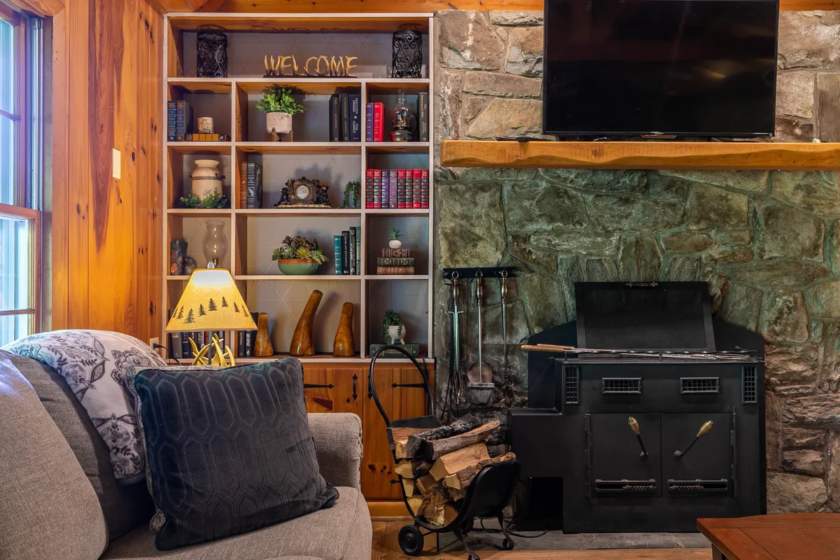 A warm, welcoming living area is depicted, centered around a stone fireplace with a bookshelf, a comfortable sofa, and soft lamp lighting. The space appears perfect for family gatherings or unwinding on a rainy day.