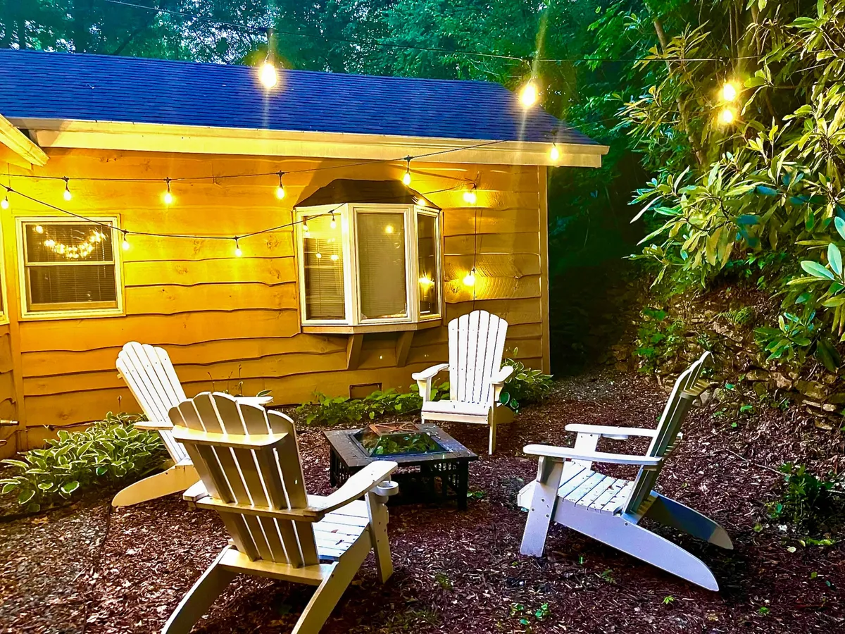 An inviting exterior scene shows a fire pit surrounded by Adirondack chairs, glowing string lights, and the house in the background. The setup encourages gatherings for s'mores, storytelling, and stargazing, evoking warmth and togetherness.