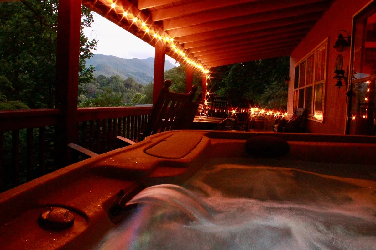 A private hot tub on the porch, twinkling string lights overhead, with a breathtaking mountain view and a cozy rocking chair nearby. The setting is ideal for both starlit evening soaks and relaxing with a drink while enjoying the scenery.