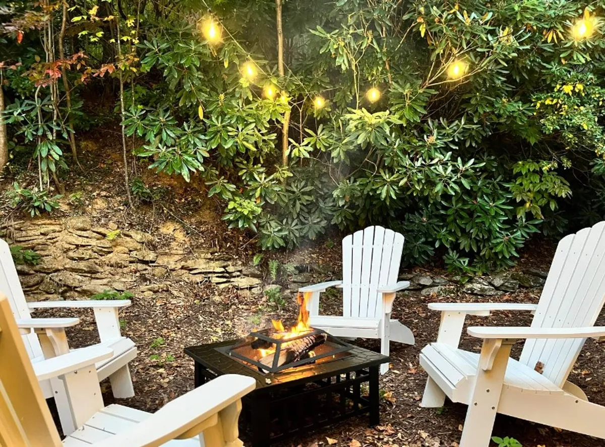 A backyard fire pit surrounded by Adirondack chairs and illuminated by charming string lights. The area is bordered by towering trees, creating a warm, communal atmosphere perfect for evening gatherings and storytelling.