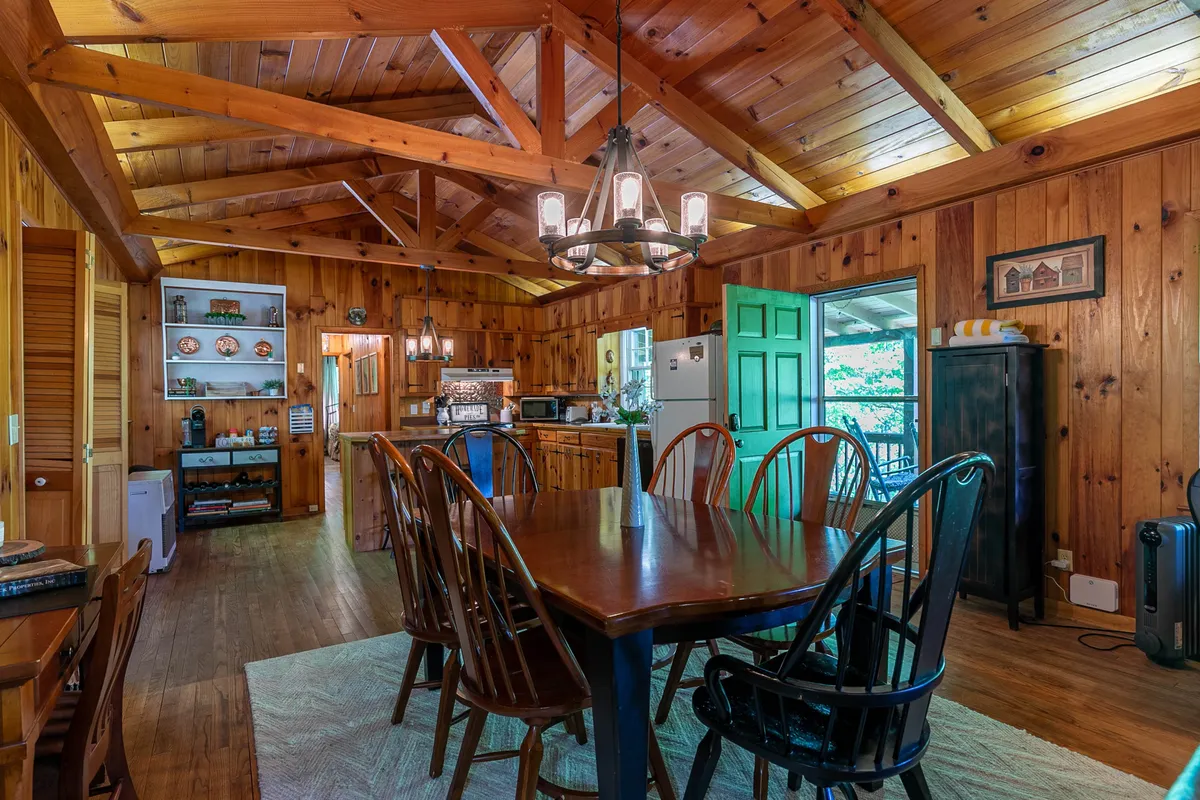 A welcoming view of the dining area and adjacent kitchen, featuring a rustic wooden table under a chandelier, exposed beams, and a glimpse of the kitchen workspace. The green door and large windows add warmth and character, suggesting an inviting communal atmosphere.