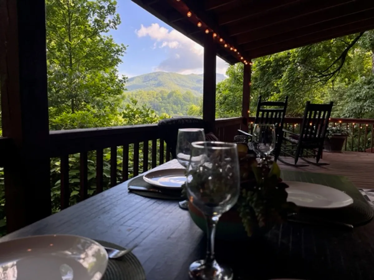 A spacious covered porch featuring a dining table set for a meal, several classic rocking chairs, and a panoramic mountain view in the background. The scene is tranquil and welcoming, ideal for both dining and relaxing outdoors.