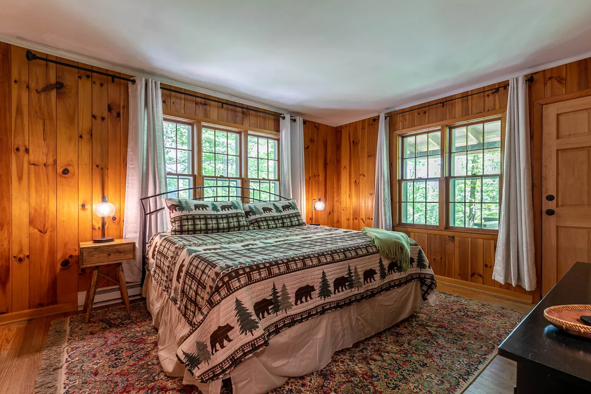 This bedroom showcases a bed with patterned bedding, rustic wooden walls, and large windows that allow natural light and tranquil nature views to pour in. The space feels serene and connected to the outdoors.