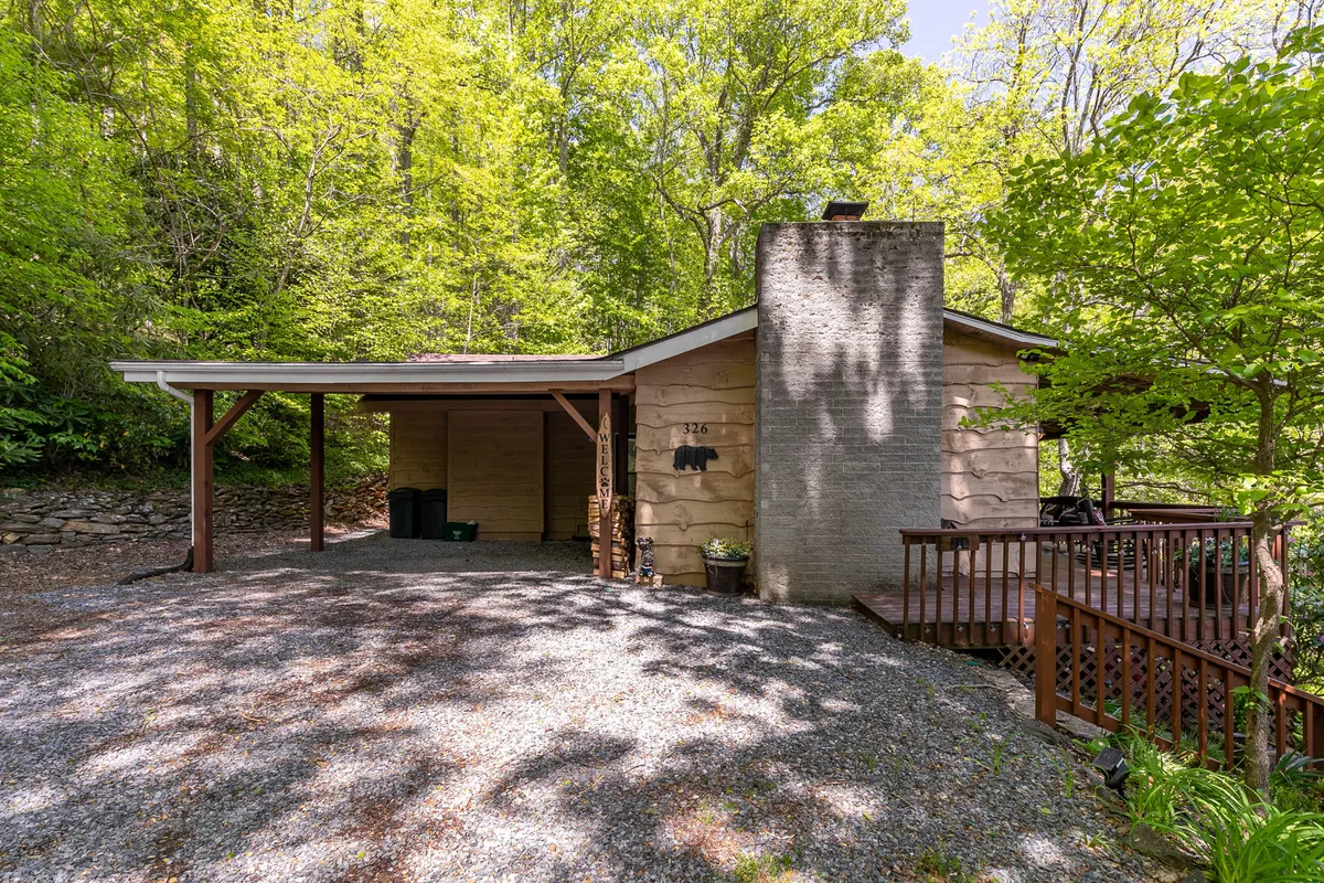 A wide wooden porch wraps around the cabin, offering inviting spaces for relaxation. The stone chimney hints at the warmth awaiting inside, while the surrounding greenery and gravel driveway immerse the cabin in a secluded, natural setting. The porch is furnished for comfort, ideal for sipping coffee or sharing a quiet moment as morning mist lifts from the nearby mountains.