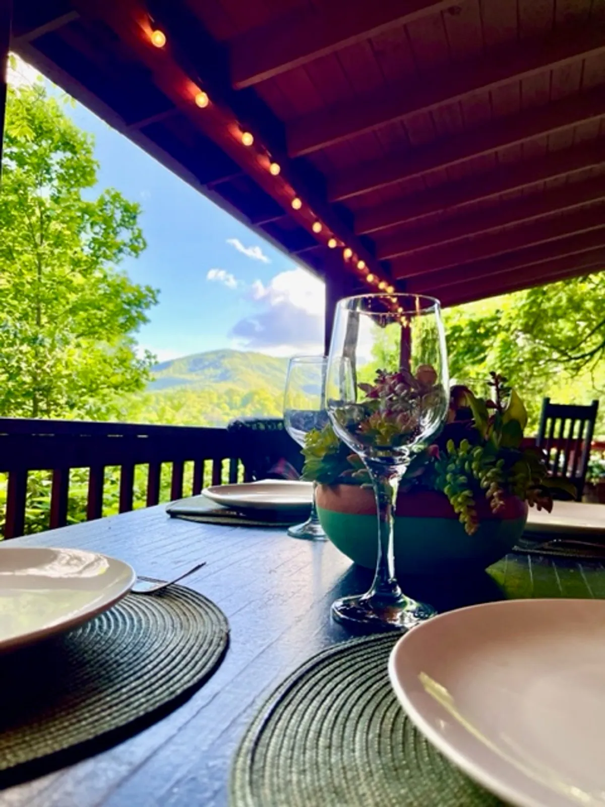 A beautifully set outdoor dining table is positioned to take full advantage of panoramic mountain views. Glasses and a decorative plant enhance the sense of a special occasion, perfect for celebrating love or sharing a quiet meal in nature's splendor.