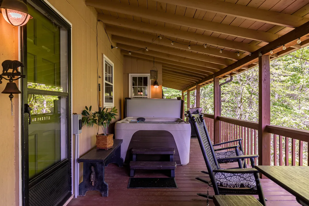 The image showcases the spacious upper deck of a rustic cabin overlooking lush greenery, featuring a bubbling hot tub, cozy outdoor seating, decorative lanterns, and vibrant plants. The serene setting invites relaxation, with sweeping views of Grandfather Mountain visible in the background.